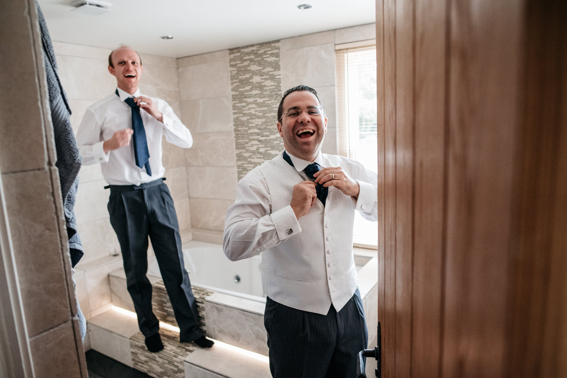 Pair of groomsmen laughing in the bathroom fixing their ties