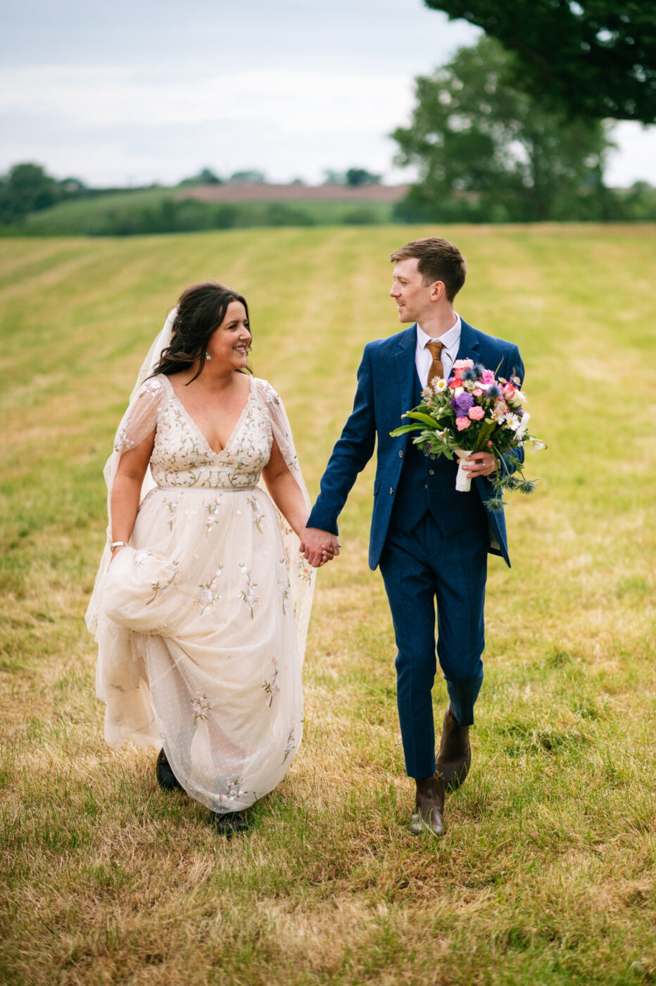 newlyweds walking through a field together with the groom holding the bouquet