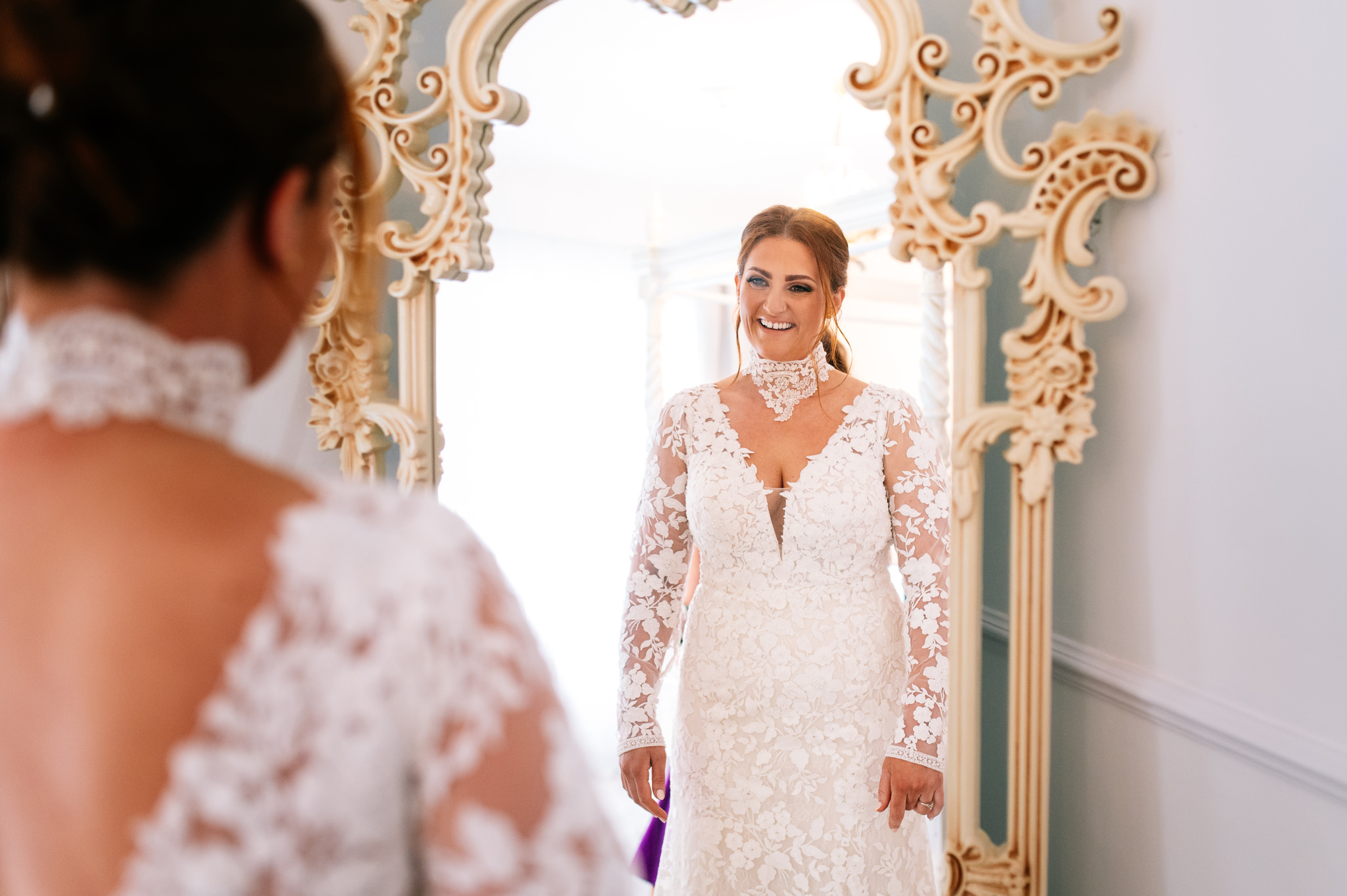 smiling bride looking at herself in the mirror wearing a wedding dress with lace collar