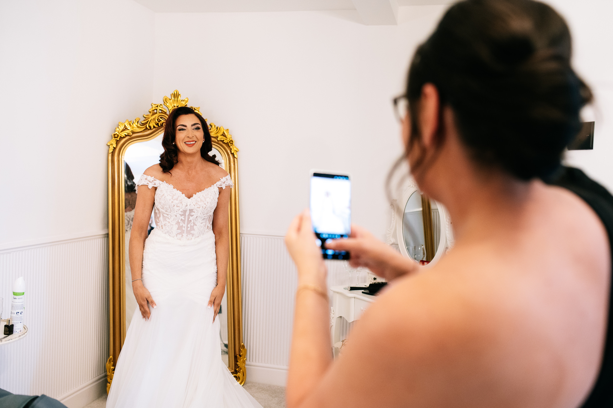 bride stood in her wedding dress in the bridal suite while a friend takes a photo