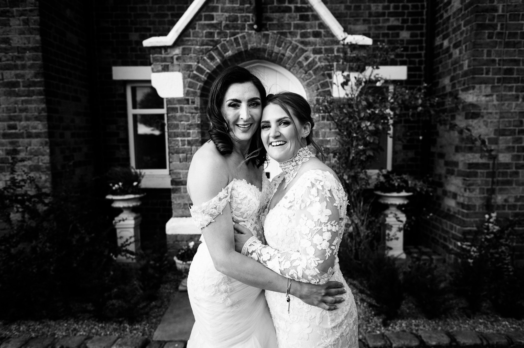 two happy brides wearing wedding dresses and hugging stood in front of the house at the cheshire venue