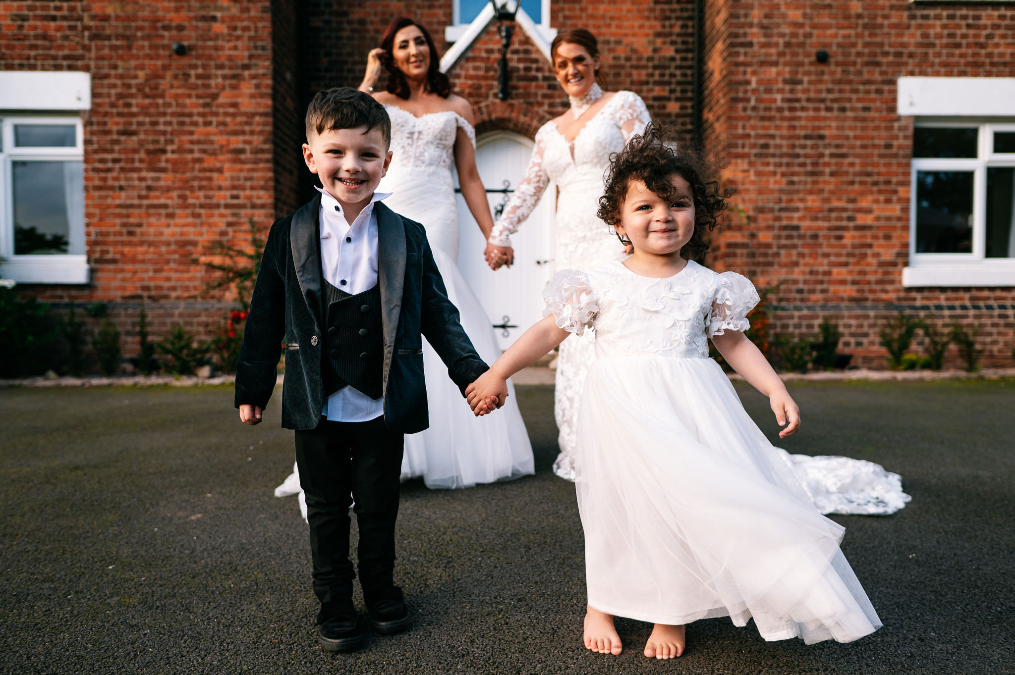 a young boy and girl in page boy and flower girl outfits with their two mums stood slightly out of focus behind them