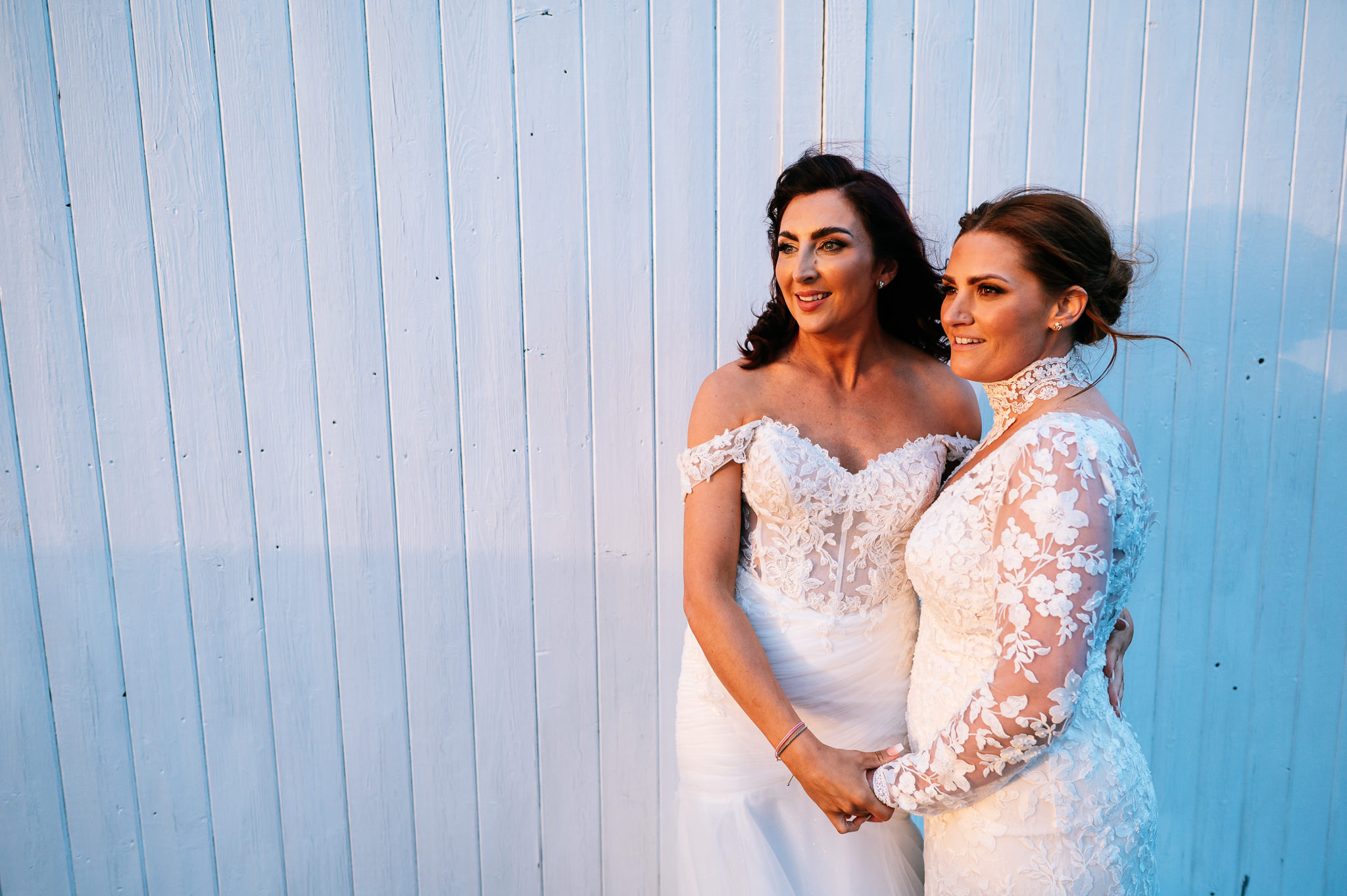 two brides stood holding hands against a white wooden door looking into the golden sunlight