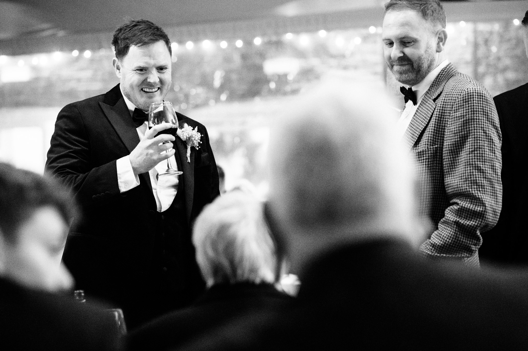 a man wearing a tuxedo holding a glass of wine and laughing with friends
