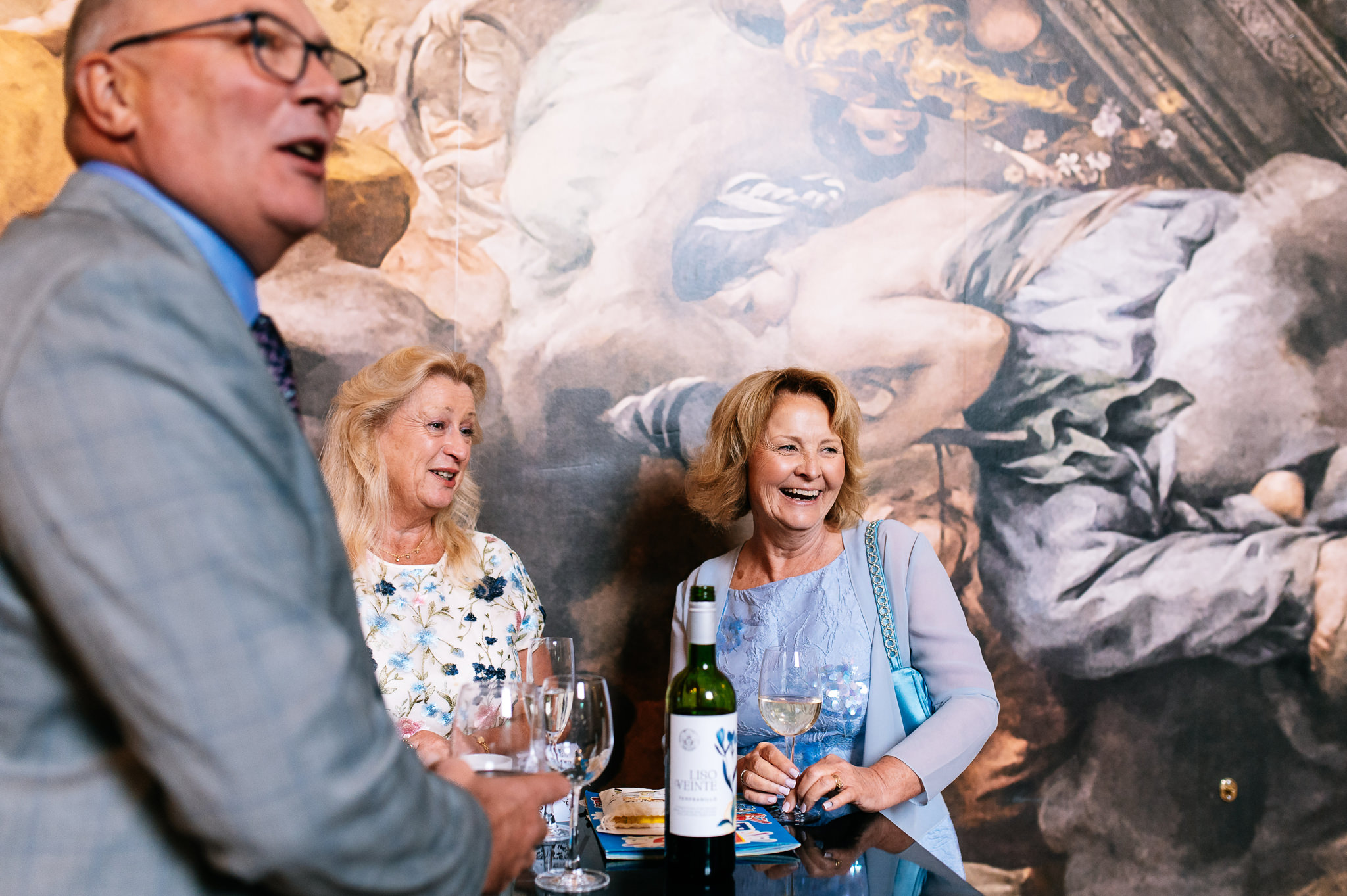two women and a man stood by a table sharing a bottle of wine and laughing together