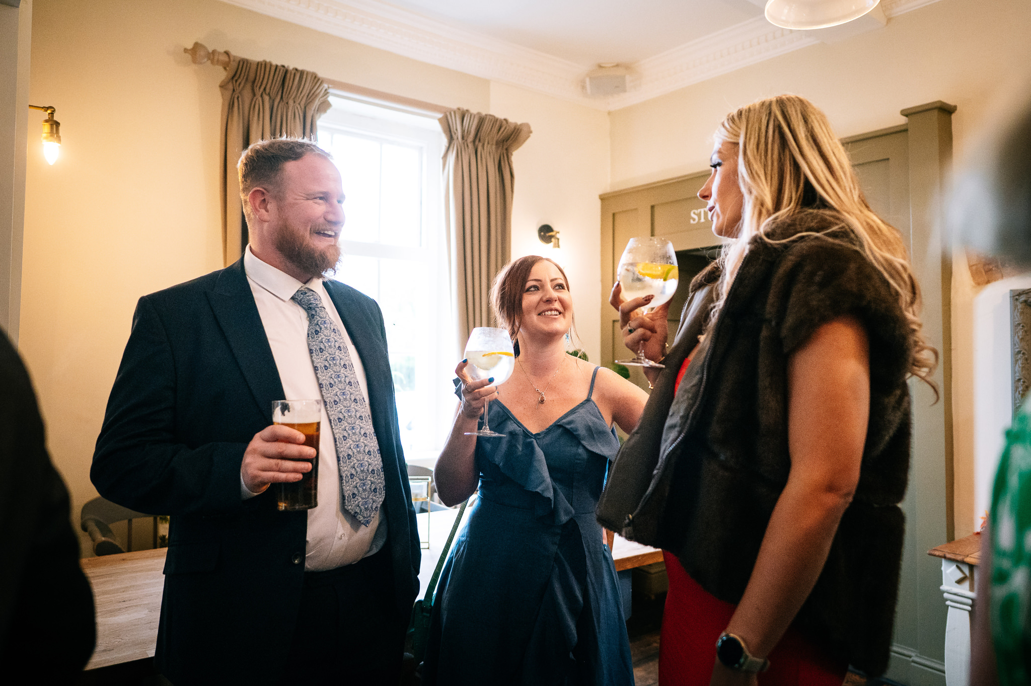 a man in a suit stood with two women in dresses, all are smiling and holding drinks
