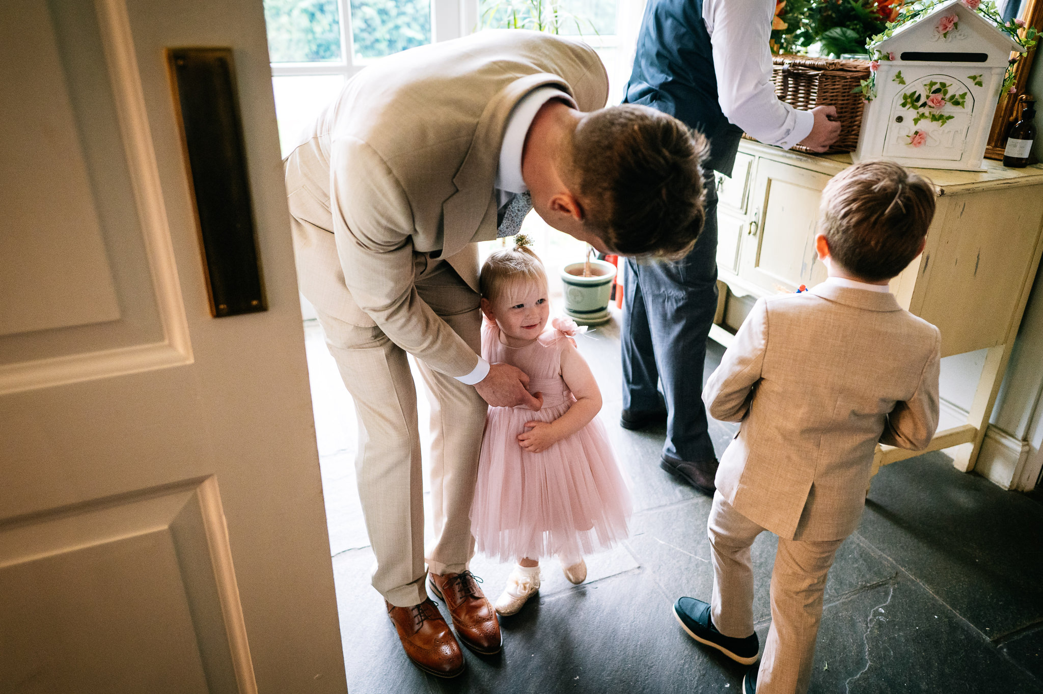 a groom wearing a light beige suit bending down to tickle his daughter who's wearing a pink dress