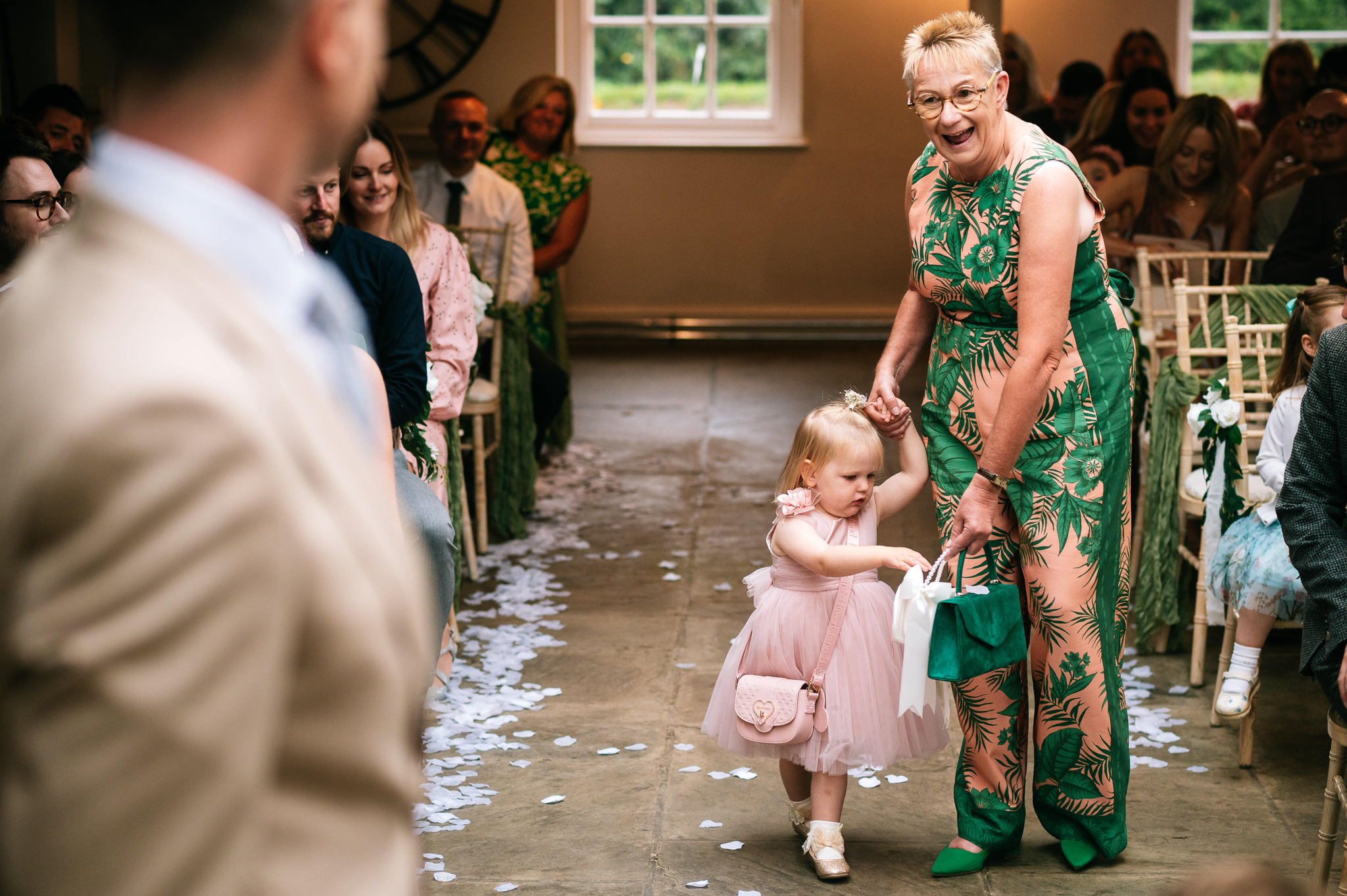 a lady in a pink and green foliage patterned jumpsuit walking down the aisle with her granddaughter who's throwing petals from a basket