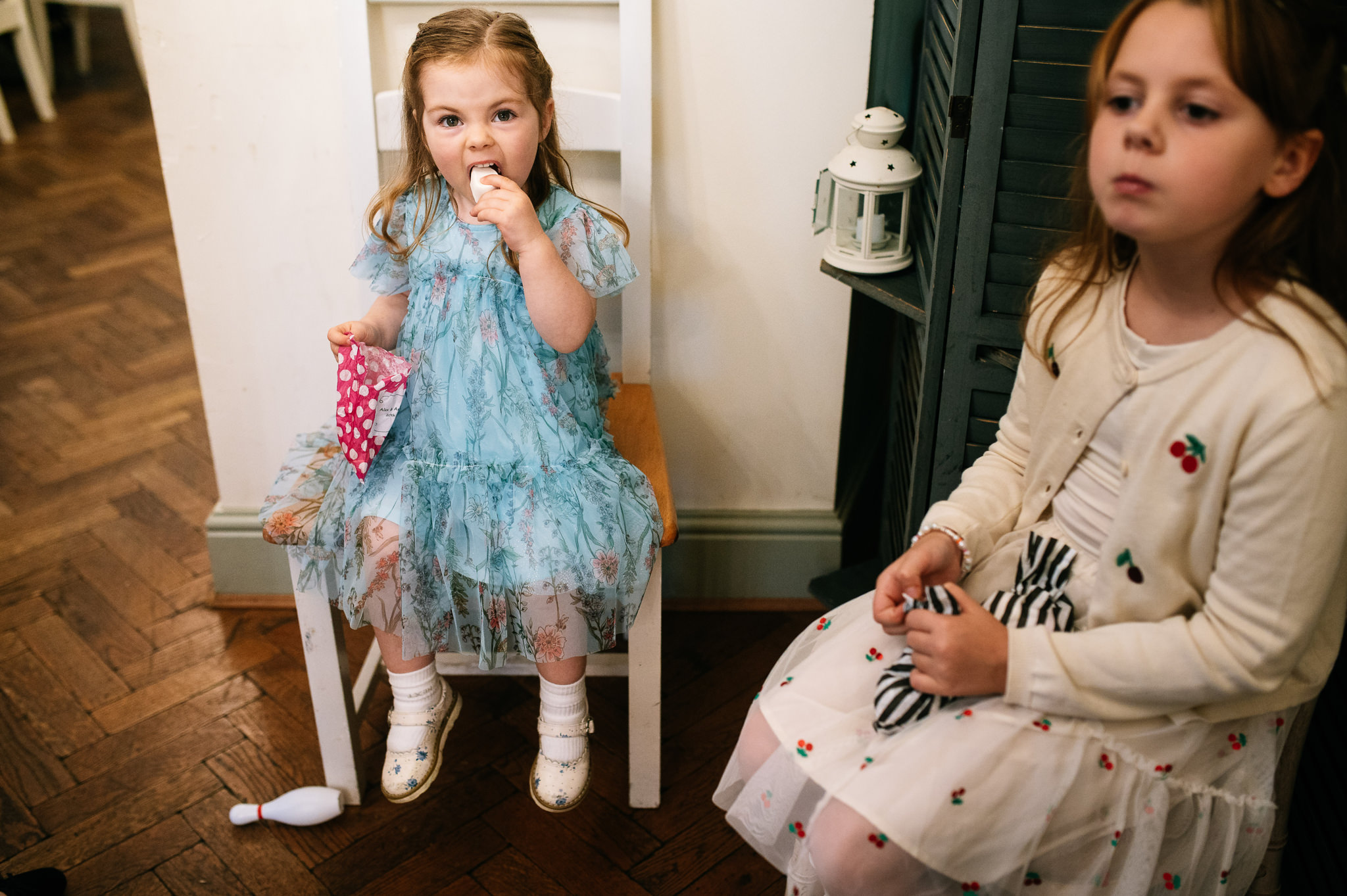 little girl in a blue dress sat on a white chair eating sweets from a paper bag with a funny expression on her face