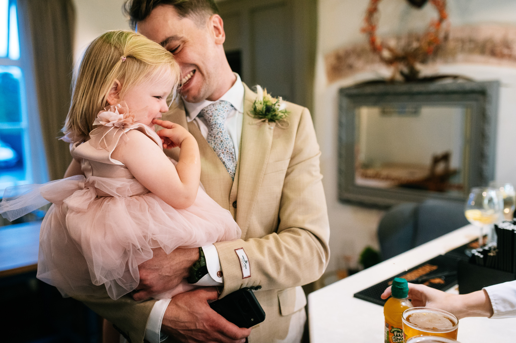 groom in a beige suit holding his young daughter and laughing whilst at the bar