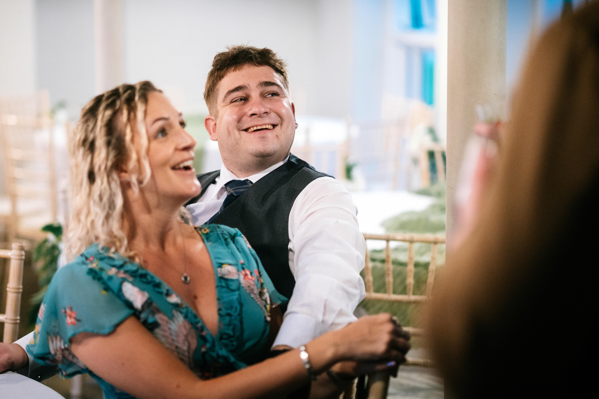 man in a white shirt and waistcoat sat at a table smiling and looking up at someone, while a woman in blue patterned dress is blurry in the foreground