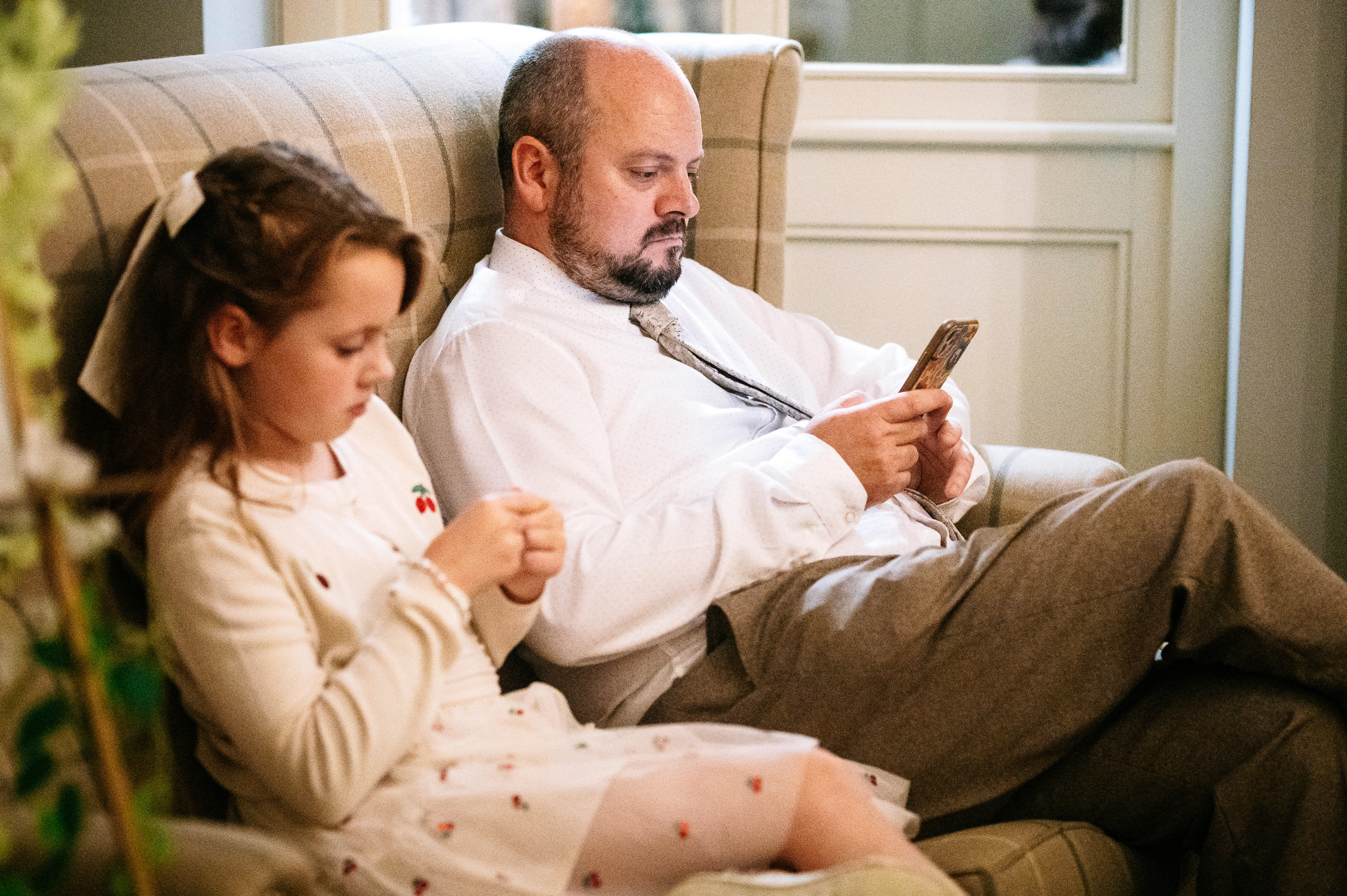 photo of a man in a shirt and tie sat on a sofa looking at his phone, while a young girl sits next to him mirroring his actions