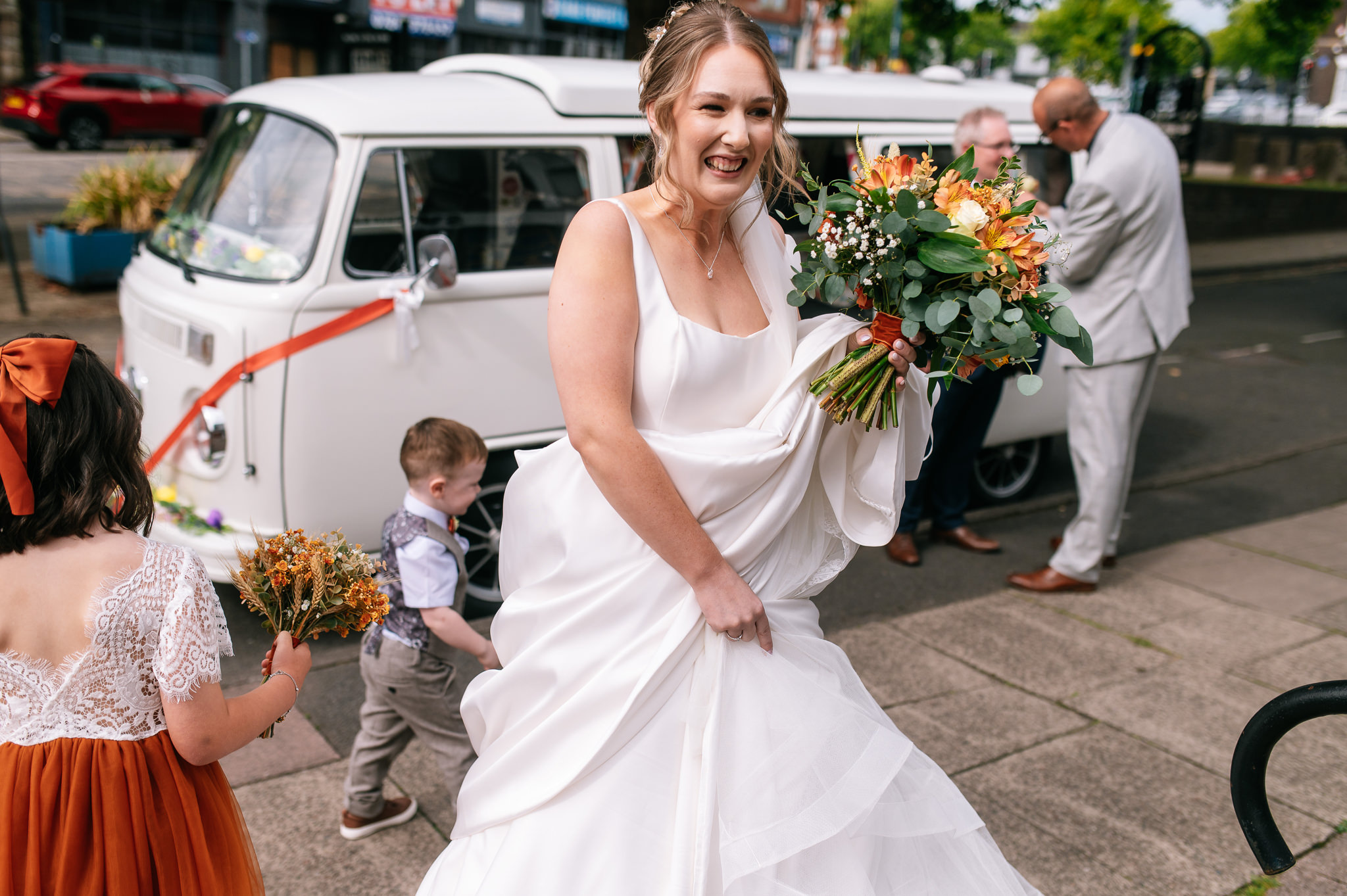 bride arriving at the church holding a rustic coloured bouquet with a white camper van behind her