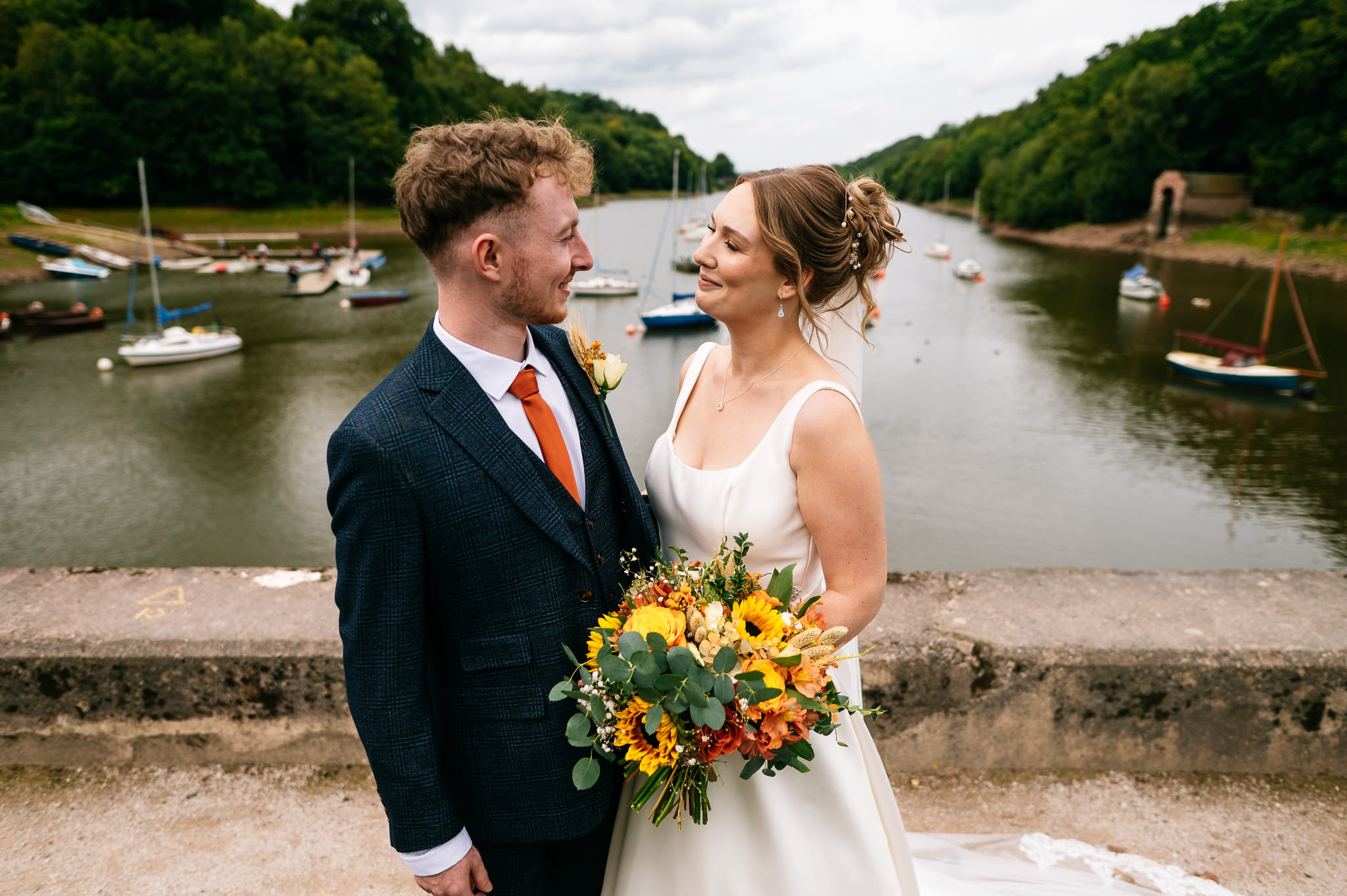 a smiling newly married couple stood in front of of rudyard lake with a number of small boats on the water