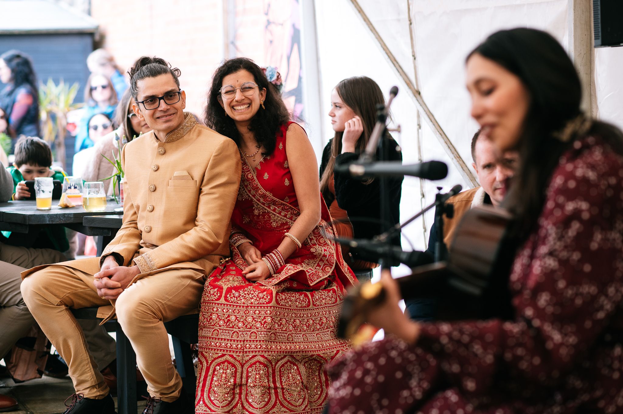 bride and groom sat together smiling and watching a lady play acoustic guitar at their wedding